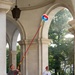Two workers are cleaning the vaulted ceiling and floor of a classical arcade or portico with white arched columns. One worker uses a long red telescoping pole with a colorful blue-and-red duster head to reach the upper ceiling, while the other holds a traditional broom. The setting appears to be an Italian public building or monument with elegant architectural arches.