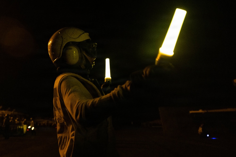 A person wearing a flight deck helmet with ear protection is holding two illuminated wands/batons in a nighttime outdoor setting. The individual appears to be a flight deck crew member performing aircraft marshalling or directing duties. The scene is consistent with nighttime flight deck operations, likely aboard a naval vessel or airfield.