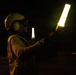 A person wearing a flight deck helmet with ear protection is holding two illuminated wands/batons in a nighttime outdoor setting. The individual appears to be a flight deck crew member performing aircraft marshalling or directing duties. The scene is consistent with nighttime flight deck operations, likely aboard a naval vessel or airfield.