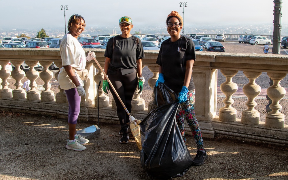 Three women are posing together during an outdoor community cleanup activity on what appears to be a terrace or promenade with a decorative stone balustrade. They are equipped with cleaning tools and protective gear, smiling at the camera. A large parking area and cityscape are visible in the background.