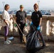 Three women are posing together during an outdoor community cleanup activity on what appears to be a terrace or promenade with a decorative stone balustrade. They are equipped with cleaning tools and protective gear, smiling at the camera. A large parking area and cityscape are visible in the background.