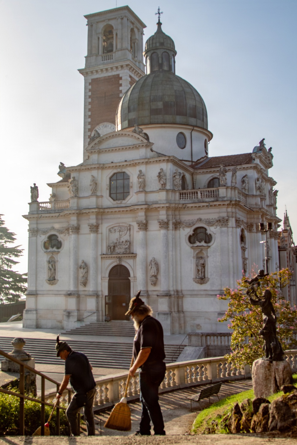 Two workers in dark uniforms and caps are sweeping a terrace or pathway in front of a large Baroque-style white marble church with a green oxidized dome and a bell tower. The church features elaborate sculptural decorations, statues in niches, and ornate facade details typical of Italian Baroque ecclesiastical architecture. The scene is captured in warm natural light, likely early morning or late afternoon.