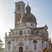 Two workers in dark uniforms and caps are sweeping a terrace or pathway in front of a large Baroque-style white marble church with a green oxidized dome and a bell tower. The church features elaborate sculptural decorations, statues in niches, and ornate facade details typical of Italian Baroque ecclesiastical architecture. The scene is captured in warm natural light, likely early morning or late afternoon.