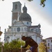 Two individuals are performing outdoor maintenance or landscaping work in a garden area in front of a large Baroque-style church or cathedral with a prominent dome and bell tower. One person is standing and using a long-handled tool (likely a shovel or rake), while the other is kneeling and holding a black garbage bag. The setting appears to be an Italian city, likely Vicenza, based on the architectural style.