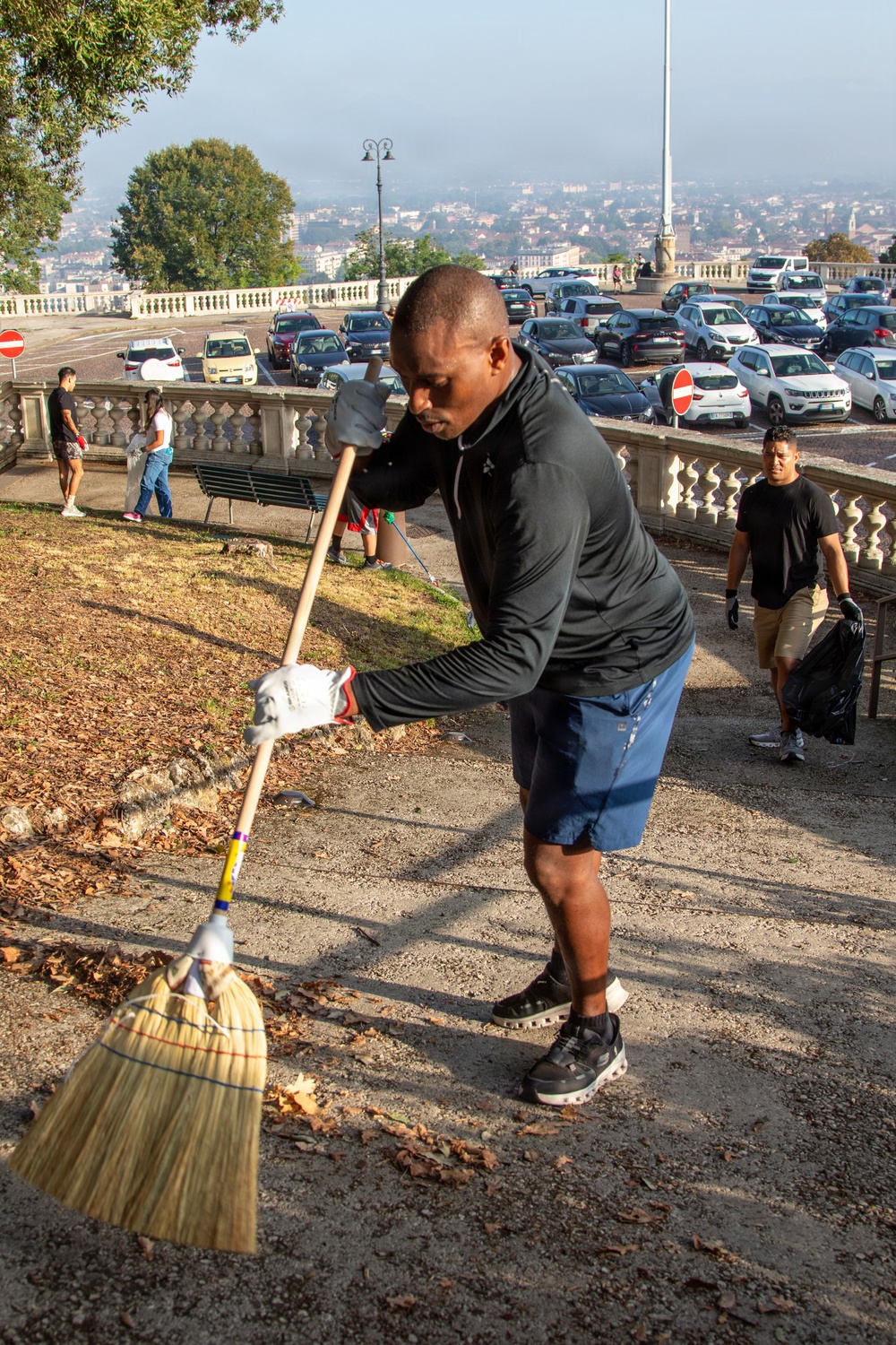 A man in a black jacket and blue shorts is actively sweeping dried leaves on a paved path using a straw broom, wearing white work gloves. The scene appears to be a community or volunteer cleanup event at an elevated terrace or promenade, with a parking lot, balustrade railing, and a cityscape visible in the background. Several other participants are visible in the background also engaged in cleanup activities.