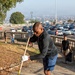 A man in a black jacket and blue shorts is actively sweeping dried leaves on a paved path using a straw broom, wearing white work gloves. The scene appears to be a community or volunteer cleanup event at an elevated terrace or promenade, with a parking lot, balustrade railing, and a cityscape visible in the background. Several other participants are visible in the background also engaged in cleanup activities.
