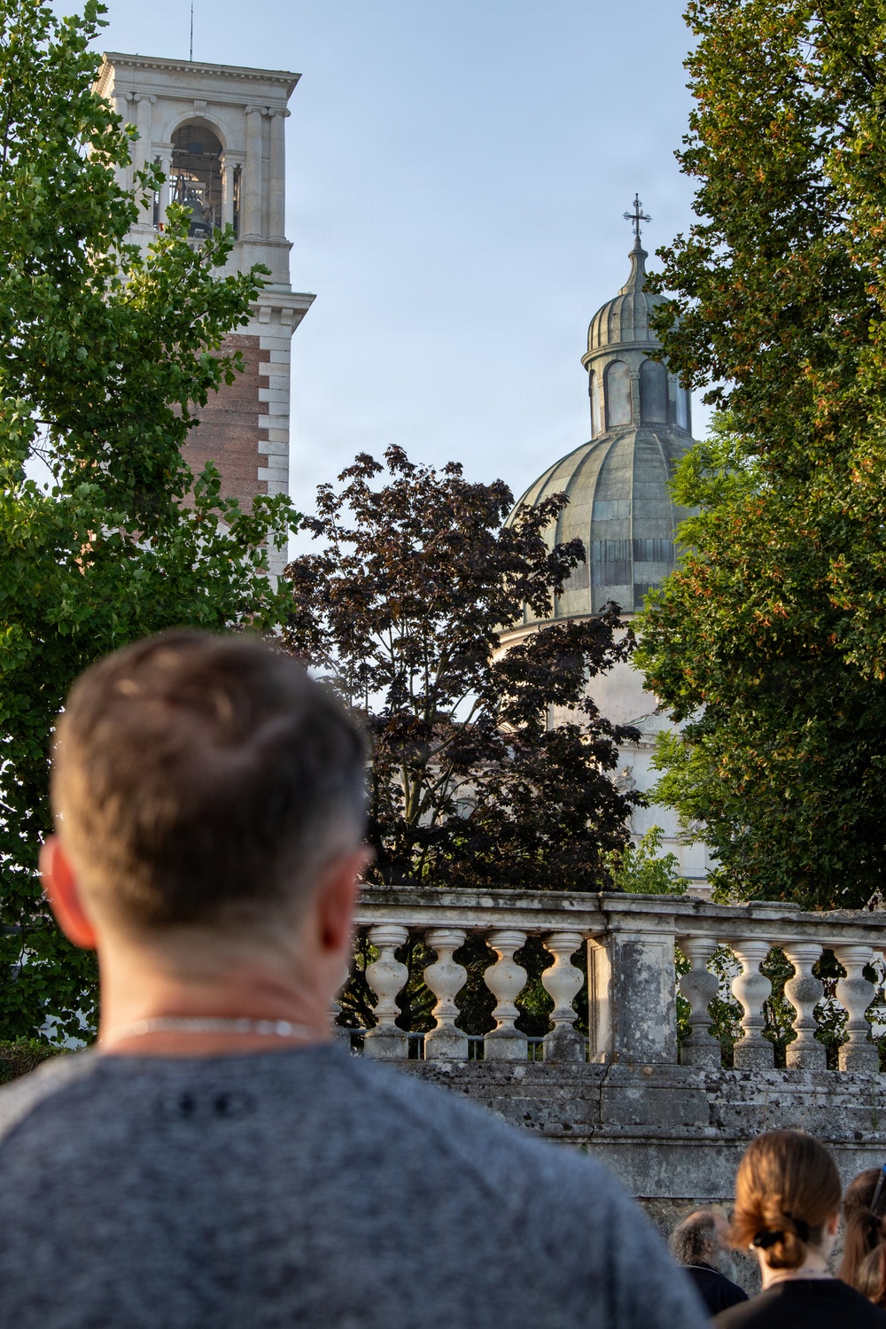 A person viewed from behind looks toward a classical European religious complex featuring a bell tower and a domed church topped with a cross. The scene is framed by lush green and dark-leafed trees, with a stone balustrade in the middle ground. The warm evening light suggests the photo was taken near sunset.