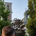 A person viewed from behind looks toward a classical European religious complex featuring a bell tower and a domed church topped with a cross. The scene is framed by lush green and dark-leafed trees, with a stone balustrade in the middle ground. The warm evening light suggests the photo was taken near sunset.