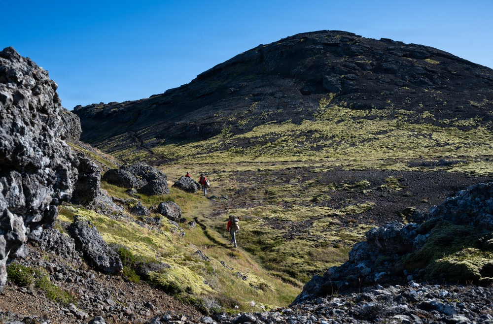 A group of hikers traverses a rugged volcanic landscape with dark basaltic rock formations and bright green moss or lichen covering the terrain. The scene appears to be in a sub-arctic or volcanic region, likely Iceland, characterized by black lava fields and sparse vegetation. Clear blue skies and strong sunlight illuminate the dramatic hillside and the small figures making their way along a trail.