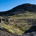 A group of hikers traverses a rugged volcanic landscape with dark basaltic rock formations and bright green moss or lichen covering the terrain. The scene appears to be in a sub-arctic or volcanic region, likely Iceland, characterized by black lava fields and sparse vegetation. Clear blue skies and strong sunlight illuminate the dramatic hillside and the small figures making their way along a trail.