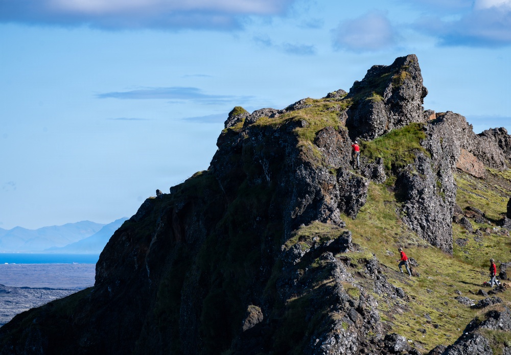 A dramatic rocky mountain ridge is shown with multiple hikers wearing red jackets navigating the rugged terrain. The landscape features dark volcanic rock partially covered with green moss and grass, with a blue ocean and distant mountains visible in the background. The scene suggests a coastal highland environment, likely in Iceland or a similar North Atlantic location.