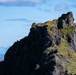 A dramatic rocky mountain ridge is shown with multiple hikers wearing red jackets navigating the rugged terrain. The landscape features dark volcanic rock partially covered with green moss and grass, with a blue ocean and distant mountains visible in the background. The scene suggests a coastal highland environment, likely in Iceland or a similar North Atlantic location.