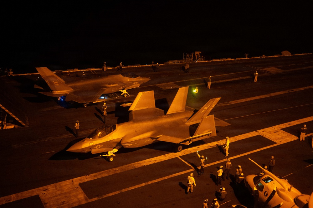A nighttime flight deck operation aboard what appears to be a U.S. Navy aircraft carrier, showing at least two F-35C Lightning II stealth fighter jets being handled by deck crew. The scene is illuminated by warm orange artificial lighting, creating dramatic contrast against the dark sky. Multiple flight deck personnel in colored jerseys are visible performing aircraft handling and marshaling duties.
