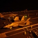 A nighttime flight deck operation aboard what appears to be a U.S. Navy aircraft carrier, showing at least two F-35C Lightning II stealth fighter jets being handled by deck crew. The scene is illuminated by warm orange artificial lighting, creating dramatic contrast against the dark sky. Multiple flight deck personnel in colored jerseys are visible performing aircraft handling and marshaling duties.