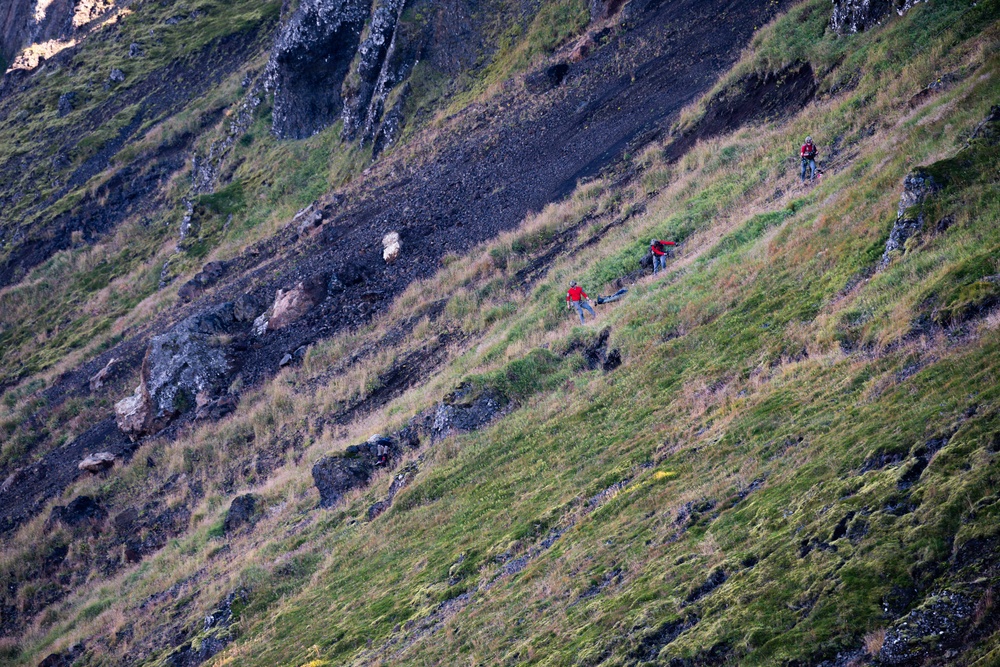 A steep grassy mountain slope with scattered dark rocky outcroppings and scree fields. Three people wearing red jackets are visible traversing the hillside, appearing to be hikers or possibly a rescue team. The terrain is rugged with a mix of green grass, dry vegetation, and dark volcanic or shale rock.