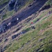 A steep grassy mountain slope with scattered dark rocky outcroppings and scree fields. Three people wearing red jackets are visible traversing the hillside, appearing to be hikers or possibly a rescue team. The terrain is rugged with a mix of green grass, dry vegetation, and dark volcanic or shale rock.
