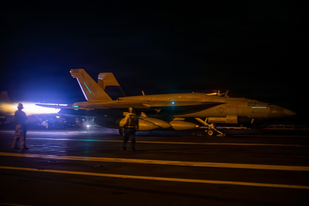 A U.S. Navy F/A-18 Super Hornet (bureau number 207) is positioned on an aircraft carrier flight deck during nighttime operations. The aircraft is illuminated by artificial lighting, with deck crew personnel visible nearby. The scene depicts pre-launch or post-recovery flight deck activity under low-light conditions.