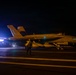 A U.S. Navy F/A-18 Super Hornet (bureau number 207) is positioned on an aircraft carrier flight deck during nighttime operations. The aircraft is illuminated by artificial lighting, with deck crew personnel visible nearby. The scene depicts pre-launch or post-recovery flight deck activity under low-light conditions.