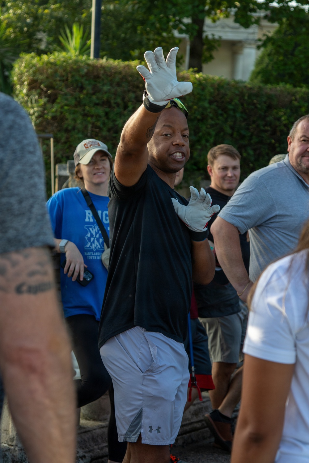 A man wearing white work gloves, a black t-shirt, and white Under Armour shorts is gesturing with both hands raised, appearing to give instructions or directions to a group of people. He is surrounded by several other individuals in an outdoor setting with green hedges and trees in the background. The scene suggests a group activity, volunteer event, or outdoor work project.