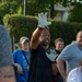 A man wearing white work gloves, a black t-shirt, and white Under Armour shorts is gesturing with both hands raised, appearing to give instructions or directions to a group of people. He is surrounded by several other individuals in an outdoor setting with green hedges and trees in the background. The scene suggests a group activity, volunteer event, or outdoor work project.