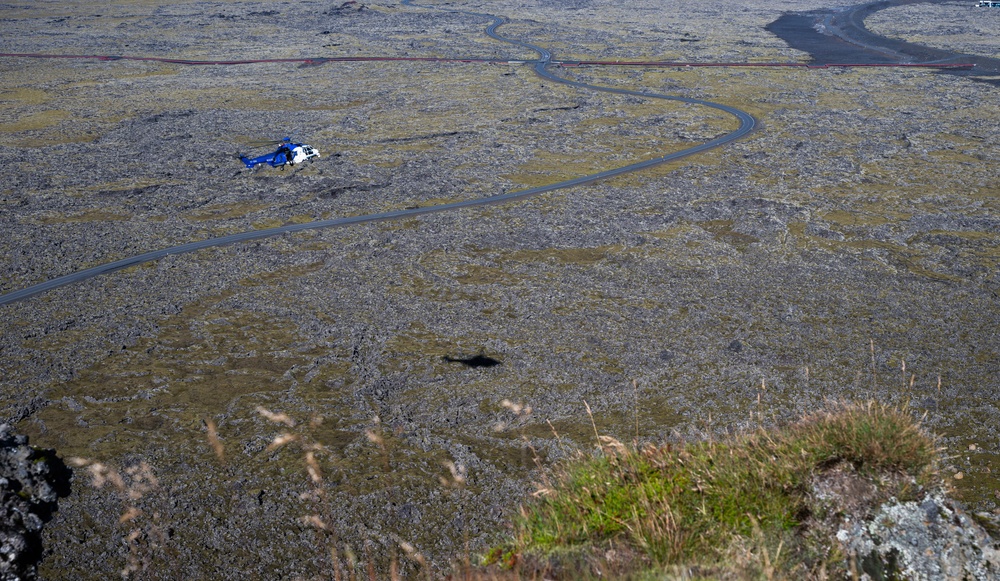 An aerial photograph taken from an elevated rocky vantage point overlooking a vast lava field landscape, likely in Iceland. A blue and white helicopter is visible flying low over the rugged terrain, which is covered in moss-covered lava rock. A winding paved road cuts through the landscape, and what appears to be a red barrier or fence line is visible in the upper portion of the image.