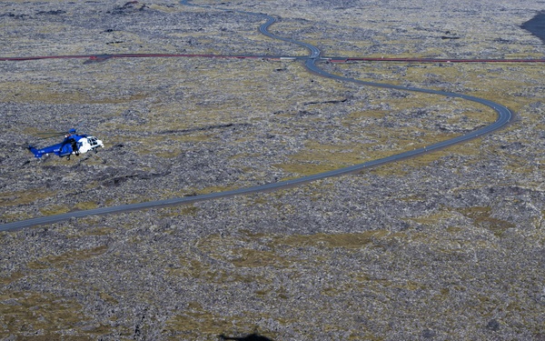 An aerial photograph taken from an elevated rocky vantage point overlooking a vast lava field landscape, likely in Iceland. A blue and white helicopter is visible flying low over the rugged terrain, which is covered in moss-covered lava rock. A winding paved road cuts through the landscape, and what appears to be a red barrier or fence line is visible in the upper portion of the image.