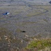 An aerial photograph taken from an elevated rocky vantage point overlooking a vast lava field landscape, likely in Iceland. A blue and white helicopter is visible flying low over the rugged terrain, which is covered in moss-covered lava rock. A winding paved road cuts through the landscape, and what appears to be a red barrier or fence line is visible in the upper portion of the image.