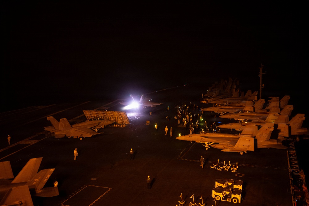 A nighttime scene on the flight deck of a naval aircraft carrier, illuminated primarily by orange deck lighting and a bright white light from what appears to be a jet blast deflector raised during a catapult launch. Multiple fixed-wing aircraft, likely F/A-18 Hornets or Super Hornets, are parked in rows along the starboard side of the deck. Numerous flight deck personnel in colored jerseys are visible moving about the deck performing various operational duties.