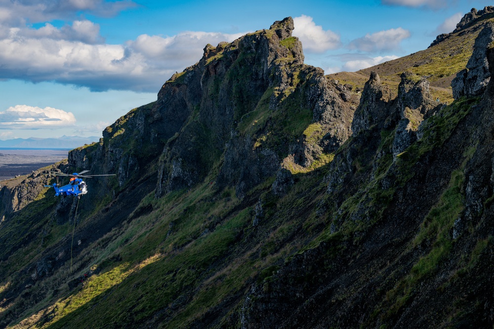 A blue rescue or coast guard helicopter hovers close to a steep, rugged mountain ridge with dark volcanic rock and green moss or grass vegetation. A cable or hoist line is visibly extended beneath the helicopter, suggesting an active rescue or winching operation on the mountainside. The background reveals a wide flat plain or coastal area under a partly cloudy blue sky.