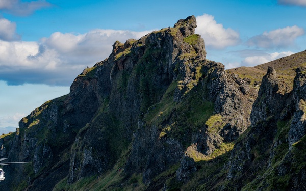 A blue rescue or coast guard helicopter hovers close to a steep, rugged mountain ridge with dark volcanic rock and green moss or grass vegetation. A cable or hoist line is visibly extended beneath the helicopter, suggesting an active rescue or winching operation on the mountainside. The background reveals a wide flat plain or coastal area under a partly cloudy blue sky.