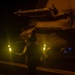 A flight deck crew member is conducting night operations on what appears to be an aircraft carrier flight deck, using illuminated marshalling wands to guide aircraft. The scene is dominated by low-light conditions with warm amber/yellow tones from the wands and ambient deck lighting. A military aircraft wing with an air-to-air missile mounted is visible overhead.