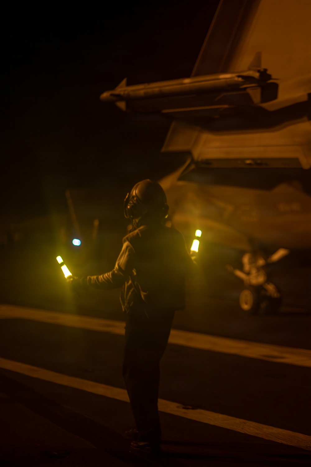 A flight deck crew member is conducting night operations on what appears to be an aircraft carrier flight deck, using illuminated wands to marshal or guide an aircraft. The scene is bathed in amber/orange light with heavy atmospheric haze, likely from jet exhaust. A military aircraft wing with an air-to-air missile mounted is visible overhead.