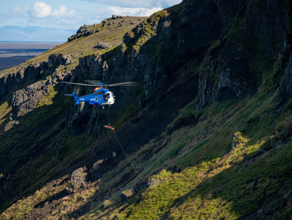A blue rescue helicopter with registration 'TF-GNA' and 'RESCUE' markings is conducting a hoist operation on a steep, rocky, moss-covered mountainside. A person or load is suspended on a cable beneath the helicopter, with additional personnel in red gear visible on the slope below. The rugged terrain appears consistent with Icelandic highland landscape.