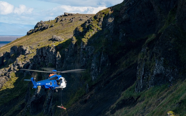 A blue rescue helicopter with registration 'TF-GNA' and 'RESCUE' markings is conducting a hoist operation on a steep, rocky, moss-covered mountainside. A person or load is suspended on a cable beneath the helicopter, with additional personnel in red gear visible on the slope below. The rugged terrain appears consistent with Icelandic highland landscape.