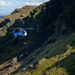 A blue rescue helicopter with registration 'TF-GNA' and 'RESCUE' markings is conducting a hoist operation on a steep, rocky, moss-covered mountainside. A person or load is suspended on a cable beneath the helicopter, with additional personnel in red gear visible on the slope below. The rugged terrain appears consistent with Icelandic highland landscape.