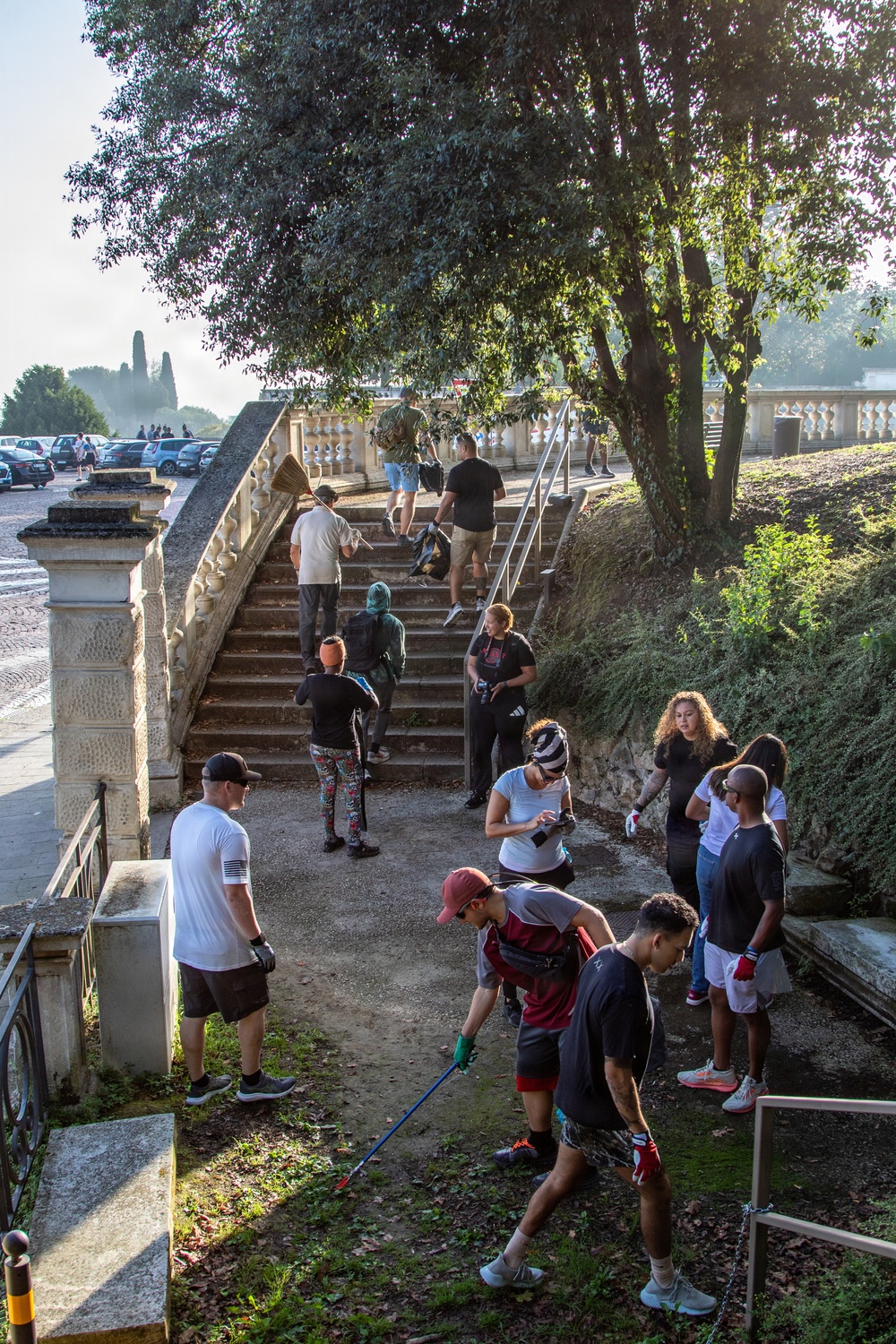 A group of approximately 12-15 people are engaged in what appears to be a community cleanup activity near an ornate stone staircase in what looks like a historic Italian park or garden. Several participants are wearing gloves and carrying litter-picking tools, collecting trash along a pathway flanked by decorative balustrades. The setting features mature trees, manicured grounds, and classical architectural elements consistent with a public heritage site.