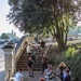 A group of approximately 12-15 people are engaged in what appears to be a community cleanup activity near an ornate stone staircase in what looks like a historic Italian park or garden. Several participants are wearing gloves and carrying litter-picking tools, collecting trash along a pathway flanked by decorative balustrades. The setting features mature trees, manicured grounds, and classical architectural elements consistent with a public heritage site.