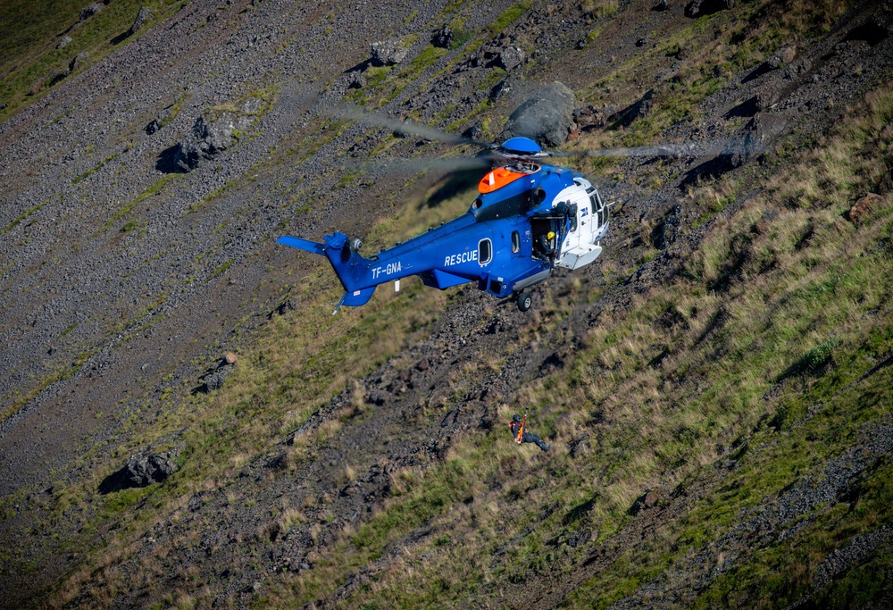 A blue and white rescue helicopter with registration TF-GNA is conducting a search and rescue operation over a rugged, rocky mountainside. A person in orange gear is visible being lowered or hoisted on a cable beneath the helicopter. The terrain consists of steep volcanic rock and sparse green vegetation, suggesting an Icelandic location.