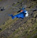A blue and white rescue helicopter with registration TF-GNA is conducting a search and rescue operation over a rugged, rocky mountainside. A person in orange gear is visible being lowered or hoisted on a cable beneath the helicopter. The terrain consists of steep volcanic rock and sparse green vegetation, suggesting an Icelandic location.