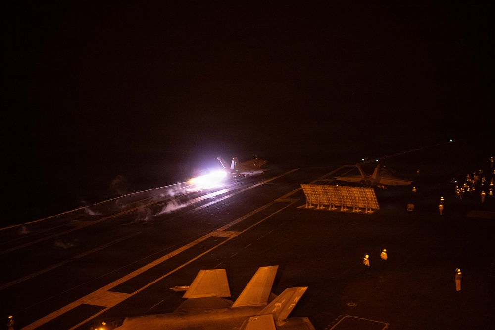 A nighttime aircraft carrier flight deck scene showing a jet aircraft launching via catapult, producing a bright engine afterburner flame and steam/exhaust trail. The image is taken from an elevated position on the carrier deck, with deck crew personnel visible as illuminated figures in the darkness. Additional parked aircraft and deck equipment are visible in the foreground and right side of the frame.