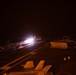 A nighttime aircraft carrier flight deck scene showing a jet aircraft launching via catapult, producing a bright engine afterburner flame and steam/exhaust trail. The image is taken from an elevated position on the carrier deck, with deck crew personnel visible as illuminated figures in the darkness. Additional parked aircraft and deck equipment are visible in the foreground and right side of the frame.