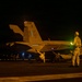 A U.S. Navy F/A-18 Hornet or Super Hornet (bureau number 202, tail code 'NF') is being prepared for a night launch on an aircraft carrier flight deck. Multiple flight deck crew members in various colored jerseys are visible performing pre-launch duties around the aircraft. The scene is illuminated by a combination of aircraft lighting, deck lighting, and ambient orange glow, consistent with nighttime carrier flight operations.