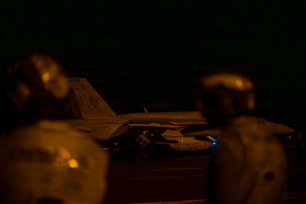 A nighttime scene on what appears to be a U.S. Navy aircraft carrier flight deck, showing at least one F/A-18 Hornet or Super Hornet jet aircraft marked with tail number '406' and 'NAVY' markings. Two personnel in flight deck gear are visible in the foreground (out of focus), while at least one crew member is visible near the aircraft. The image is taken in low-light conditions with warm amber tones from deck lighting.