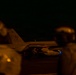 A nighttime scene on what appears to be a U.S. Navy aircraft carrier flight deck, showing at least one F/A-18 Hornet or Super Hornet jet aircraft marked with tail number '406' and 'NAVY' markings. Two personnel in flight deck gear are visible in the foreground (out of focus), while at least one crew member is visible near the aircraft. The image is taken in low-light conditions with warm amber tones from deck lighting.