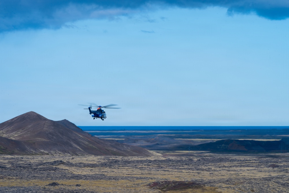 A military or search-and-rescue helicopter is flying low over a stark, volcanic highland landscape with dark reddish-brown hills and sparse vegetation. The terrain appears barren and remote, consistent with subarctic or volcanic regions such as Iceland. A large body of water or ocean is visible on the horizon under a partly cloudy blue sky.