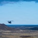 A military or search-and-rescue helicopter is flying low over a stark, volcanic highland landscape with dark reddish-brown hills and sparse vegetation. The terrain appears barren and remote, consistent with subarctic or volcanic regions such as Iceland. A large body of water or ocean is visible on the horizon under a partly cloudy blue sky.