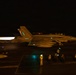 A military jet aircraft, consistent with an F/A-18 Hornet or Super Hornet, is captured during a night launch from what appears to be an aircraft carrier flight deck. The aircraft's engines are producing visible afterburner exhaust glow on the left side of the frame. Deck crew personnel and flight deck lighting are visible in the foreground and background.