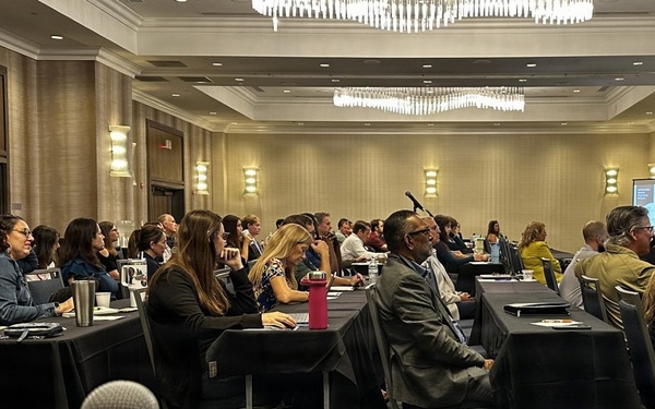 A professional conference or seminar is taking place in a hotel ballroom, with approximately 40-50 attendees seated at black-draped tables facing a presenter at the front of the room. A female speaker stands at a podium near a projection screen on the right side of the room. The event appears to be actively recorded or livestreamed, as evidenced by a laptop and microphone visible in the foreground.
