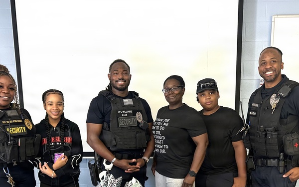 A group of six individuals, including security personnel and police officers, pose together at what appears to be a school supply drive or community outreach event. They are standing behind tables filled with backpacks, blue drawstring bags, and boxes containing school supplies. An Amazon Prime box with the phrase 'Without you, I'm empty inside' is prominently visible in the foreground.