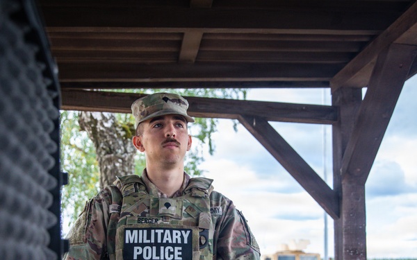 A U.S. Army Military Police soldier, identified by the name tape 'Berger,' stands at what appears to be a checkpoint or guard post wearing full tactical gear including a plate carrier vest with 'MILITARY POLICE' markings. The soldier is in OCP (Operational Camouflage Pattern) uniform and appears to be on duty at an outdoor installation, with a military vehicle (HMMWV/Humvee) visible in the background. The setting suggests a forward operating base or training area in a temperate region.